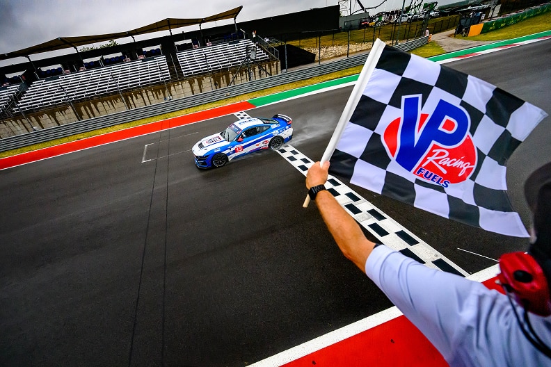 A blue and white race car with the number 13 crosses a checkered finish line on a race track